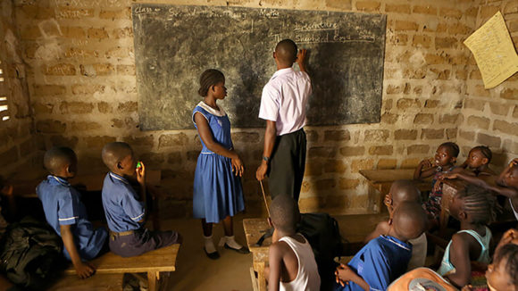 Et skarpt bilde av et klasserom i Sierra Leone. Læreren står foran tavla sammen med en elev mens de andre klassekameratene ser på.