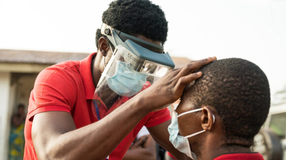 En øyehelsearbeider iført maske og visir sjekker en manns øyne for tegn på trakom.