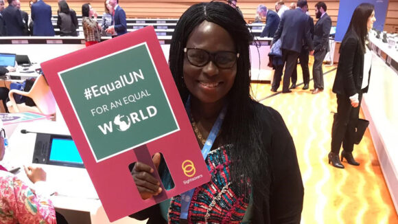 Gertrude Oforiwa Fefoame smiler og holder opp en plakat som sier '#EqualUN for en likestilt verden'.