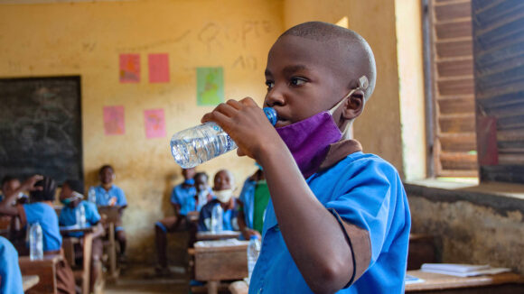 En skolegutt i Kamerun drikker vann i et klasserom etter å ha tatt ormekurmedisin.