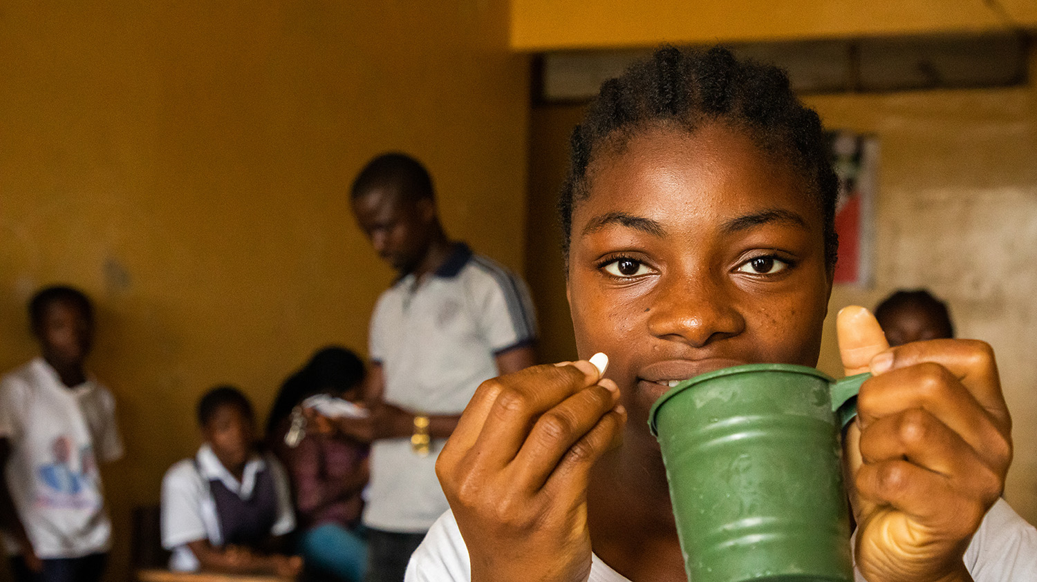 En jente på skolen holder en grønn kopp og et ormenettbrett, med klassekameratene i bakgrunnen.