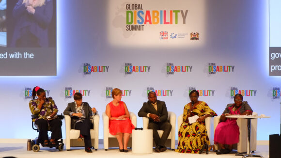 Et panel med seks talere sitter på scenen under Global Disability Summit i 2018.