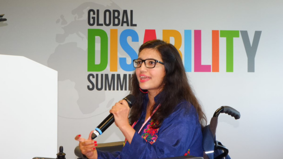 Funksjonsaktivist Abia Akram snakker i mikrofon fra rullestolen sin på scenen under Global Disability Summit