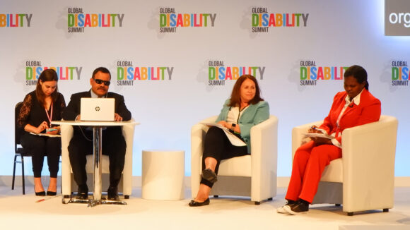 Publikumsmedlemmer under Global Disability Summit 2018 med ryggen mot kameraet, ser på skjermer med teksten 