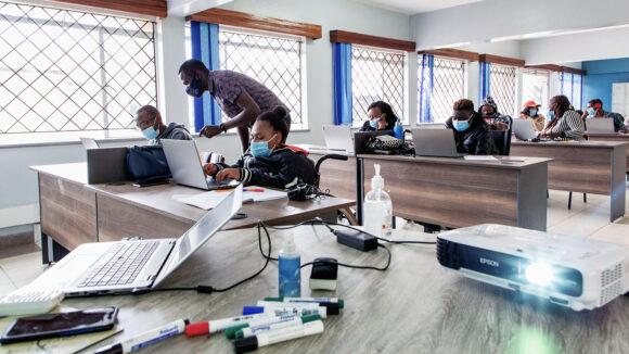 Elevene sitter foran bærbare datamaskiner ved pulter i et klasserom. I forgrunnen står en lærer ved siden av en elev og gir instruksjoner.