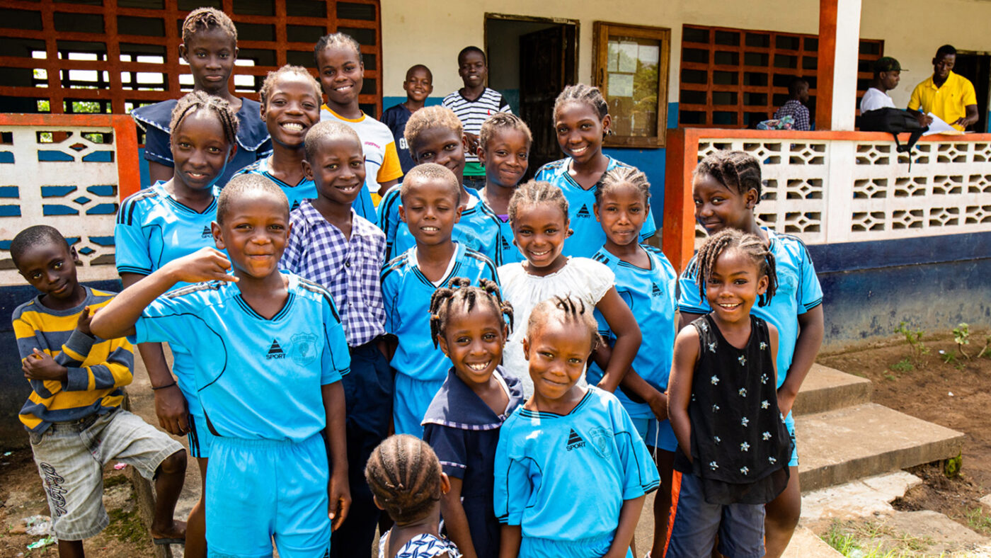 En stor gruppe smilende barn samles til gruppebilde utenfor skolen sin.
