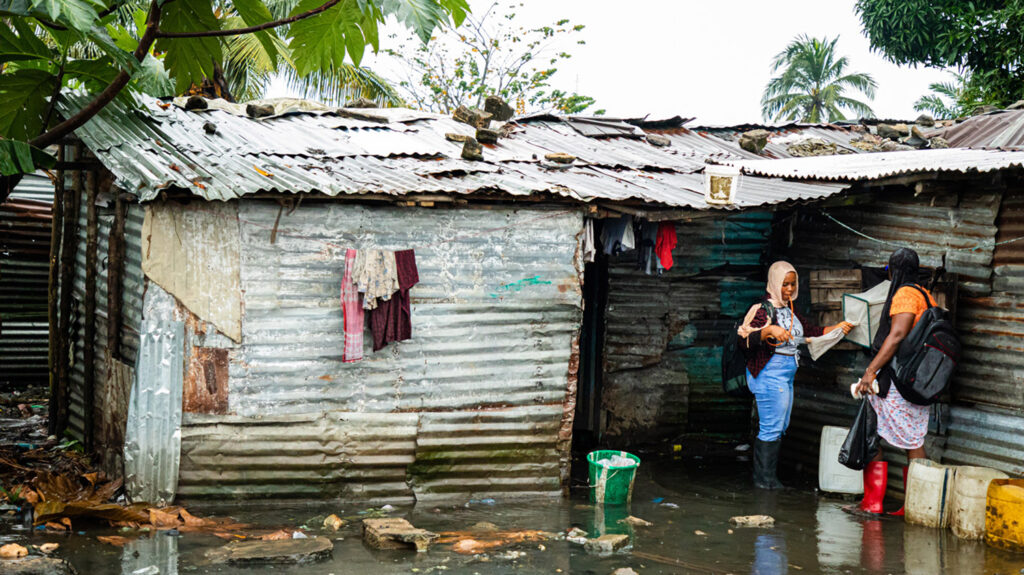 To kvinner står i et oversvømt område utenfor et hus, hvor de sjekker en myggfelle.
