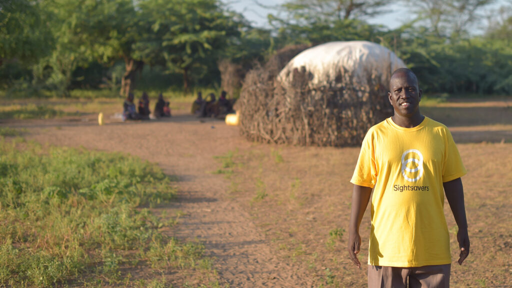 Samson står i et ørkenmiljø, iført en gul Sightsavers-t-skjorte. I bakgrunnen sitter en gruppe mennesker i ring utenfor et telt.