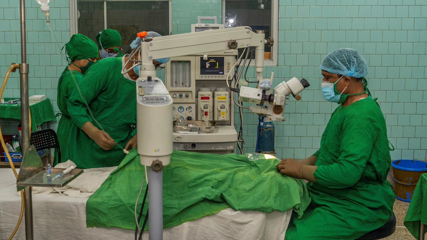 An operation takes place with a machine being looked through by a surgeon. There are four staff members wearing green scrubs, masks and headwear.