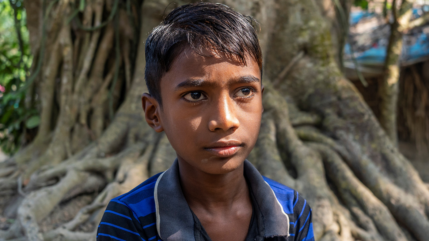 A young boy, Shahadeb, stands outside looking past the camera in sunlight.