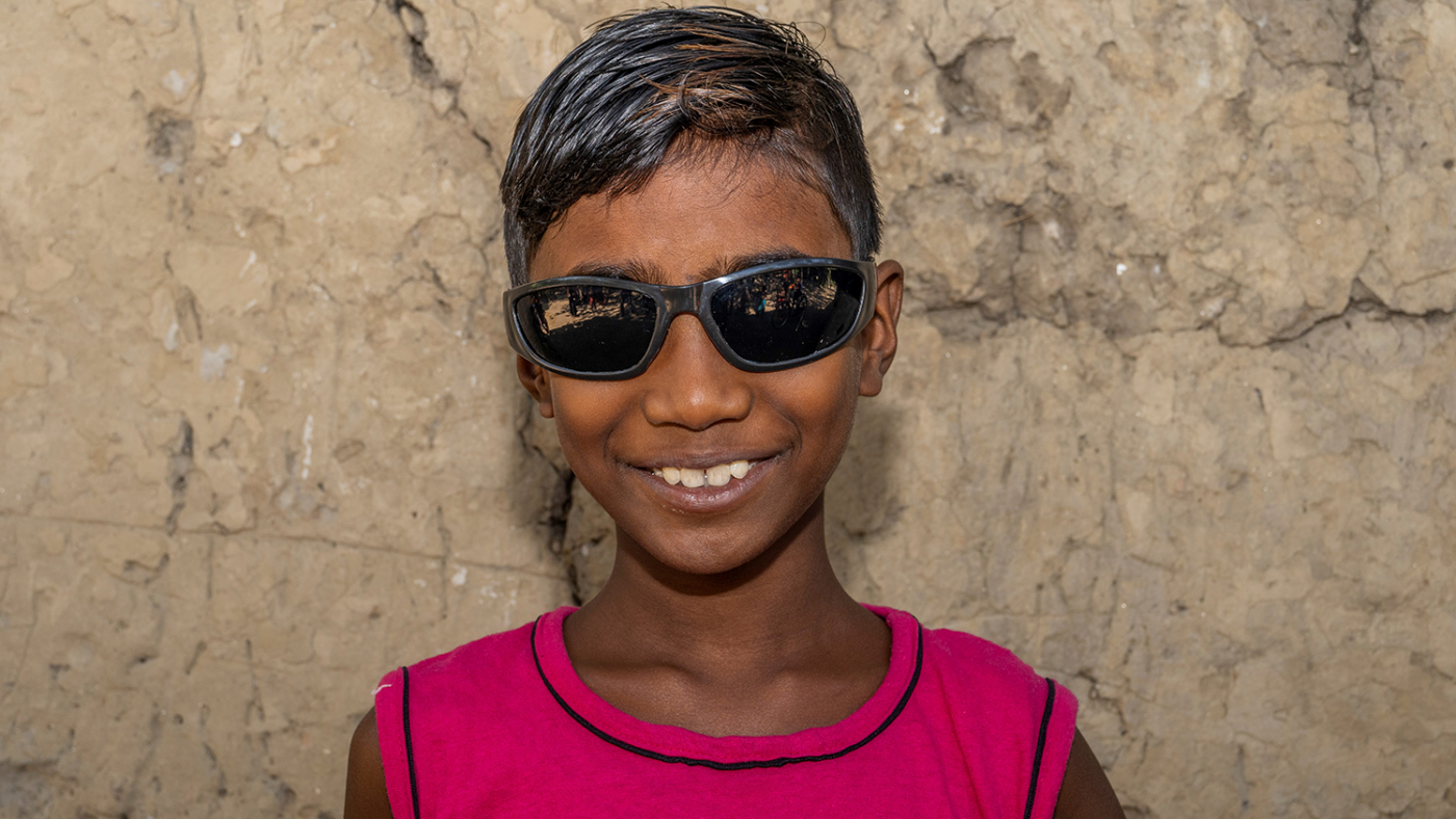 A young boy, Shahadeb, smiles widely at the camera outside wearing sunglasses.