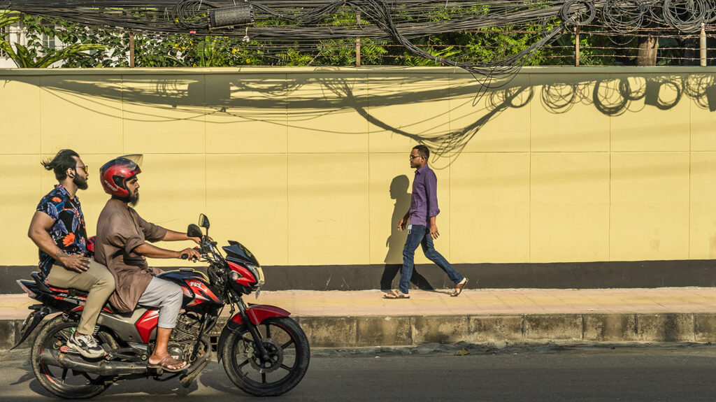 Arif går nedover en urban gate mens en motorsykkel med to førere passerer forbi på veien.