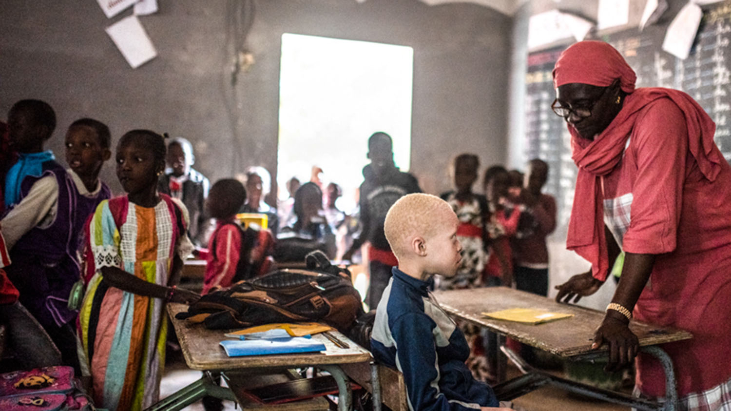 En lærer gir veiledning til en ung gutt med albinisme i et travelt klasserom i Senegal.