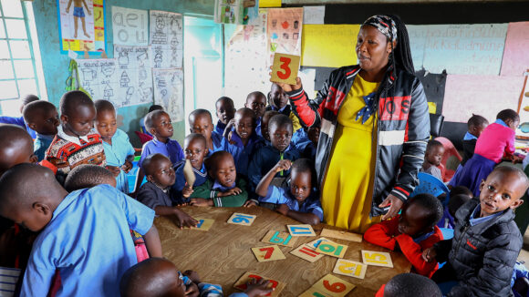 Et klasserom i Kenya, der en lærer viser et flashkort med tallet tre på til elever som sitter rundt et bord.