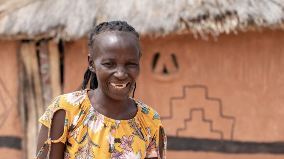 Maria smiles after her successful operation for advanced trachoma. She's standing outside her home, which has a thatched roof.