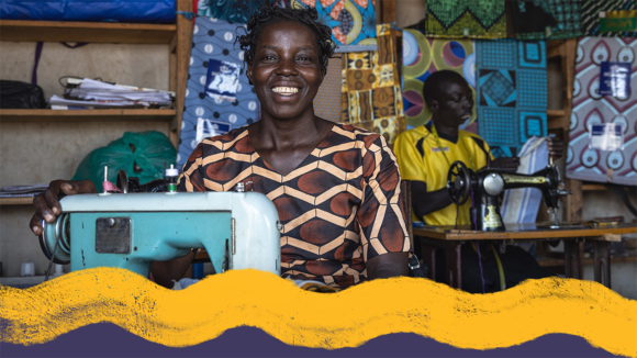 En kvinne smiler bredt mens hun sitter ved en symaskin. Bak henne inspiserer en mann et stoffstykke.