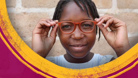 En jente med røde briller står foran en murvegg.