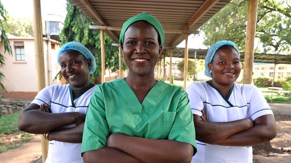 Dr. Gladys Atto, iført operasjonsklær, står sammen med to kolleger utenfor et sykehus i Uganda. De står med armene i kors og smiler mot kameraet.