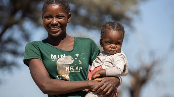 Lupula is wearing a green t-shirt and holding Sarah, aged 17 months, on her hip. Sarah is wearing a white cardigan and looking slightly off to the side.
