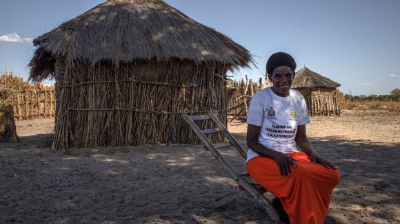 Zita sits outside in a rural village in Zambia. She wears a white t-shirt and red skirt.