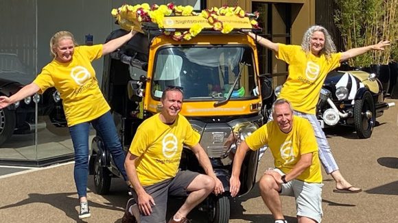 Fire innsamlere, iført gule T-skjorter med Sightsavers-logo, poserer ved siden av en tuk-tuk.